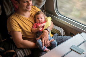 Un papa souriant tenant un bébé dans ses bras, tous deux à l'intérieur d'un train, en route vers un camping familial. L'image évoque un moment chaleureux et joyeux en famille, illustrant l'aventure et la connexion entre générations. Les voyages en famille, les vacances au camping et les expériences en train avec des enfants.